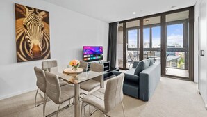 A modern living room with a glass dining table, grey upholstered chairs, a grey sofa, a mounted TV, and a large zebra painting on the wall.