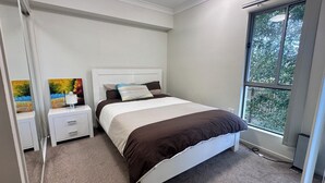 Un dormitorio con cama, mesitas de noche, ventilador de techo y ventana con vista a árboles.