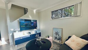 Una sala de estar con un televisor de pantalla plana sobre un mueble blanco, una mesa de centro negra, un sofá oscuro y una alfombra estampada.