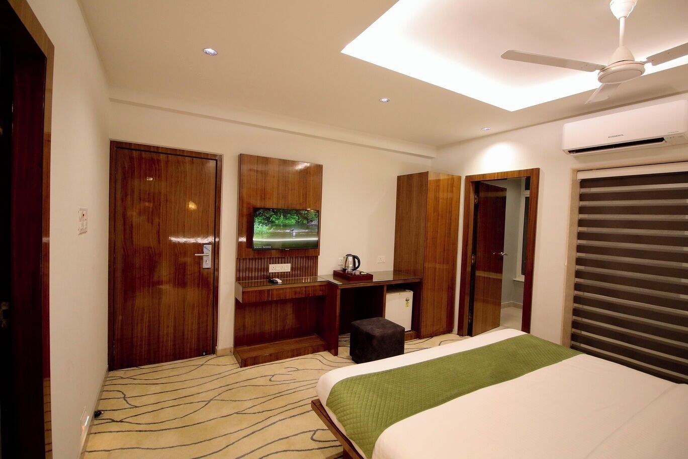 A hotel room with a bed, a desk with a television, a wardrobe, and an air conditioning unit.