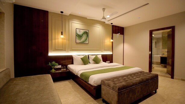 A modern bedroom with a large bed, a bedside table with a lamp, a ceiling fan, and a view of a bathroom. - Evara By Meraki Inn (Kolkata)