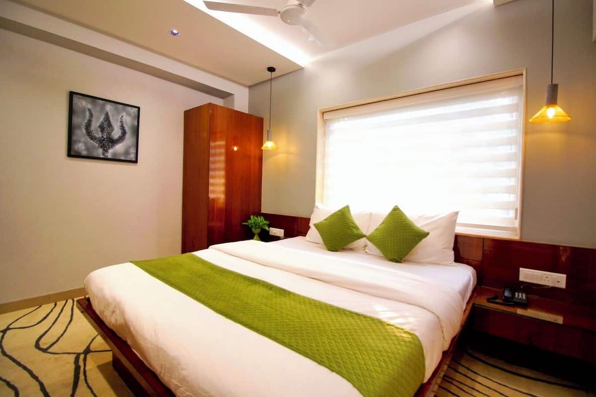 A neatly made bed with a green throw, a wooden wardrobe, a wall-mounted picture, and a window with blinds.