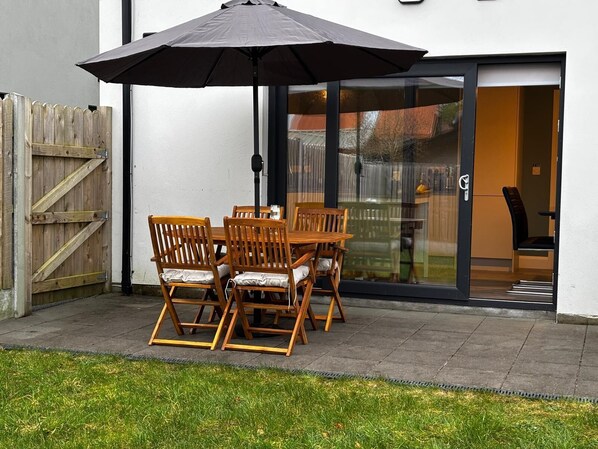 Een terras met een houten tafel en stoelen, een zwarte parasol en een glazen deur naar een binnenruimte.