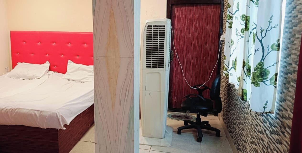 A bedroom with a red headboard, a white bedspread, a standing heater, a black office chair, and a patterned curtain.