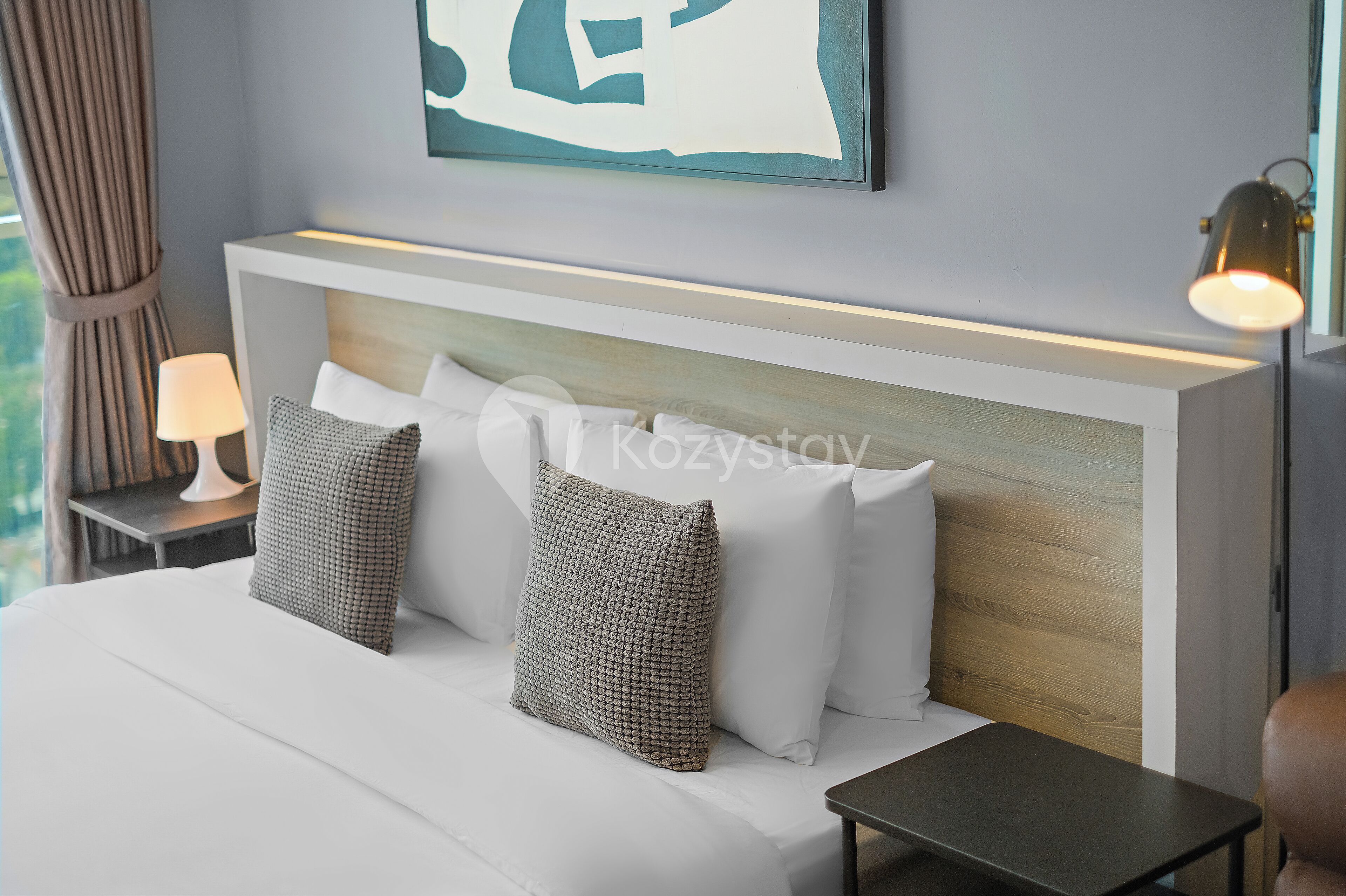 A neatly arranged hotel room with a bed, two pillows, a bedside table with a lamp, and a wall-mounted artwork.