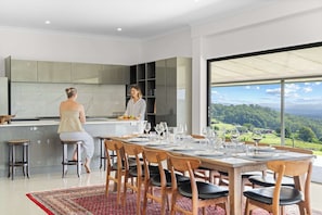 A modern kitchen with a large island, a dining area with wooden chairs, and a view of a green landscape through large windows. - Panorama Farm - Sunshine Coast Hinterland (Hinterland Region)