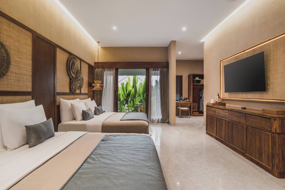A hotel room with two beds, a TV, a wooden reception desk, and a view of greenery through the window.