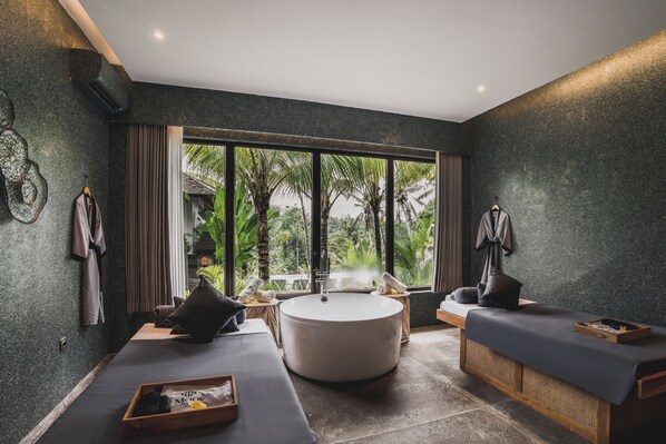 A modern spa room with a large round bathtub, two massage tables, and a view of palm trees through large windows. - Maar Resort Ubud by Ini Vie Hospitality (Payangan)