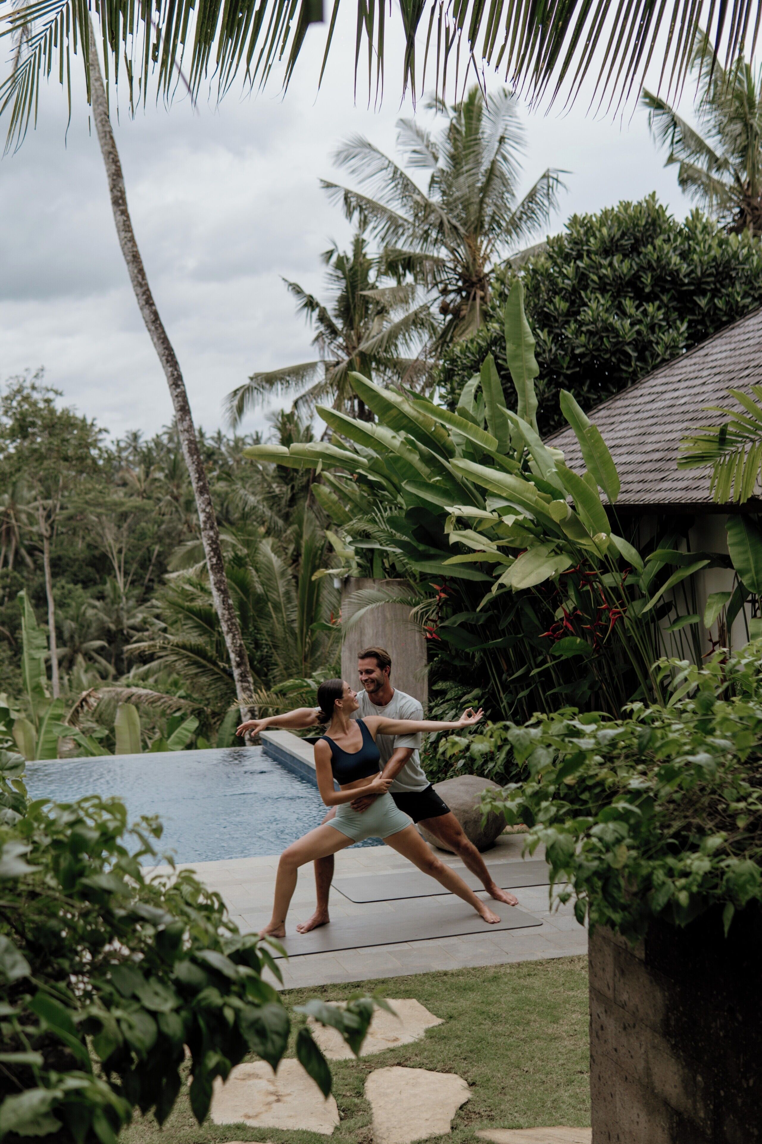 Una pareja junto a una piscina, con vegetación tropical y una casa al fondo.