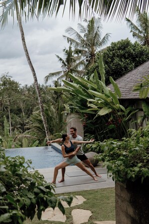 Una pareja junto a una piscina, con vegetación tropical y una casa al fondo.
