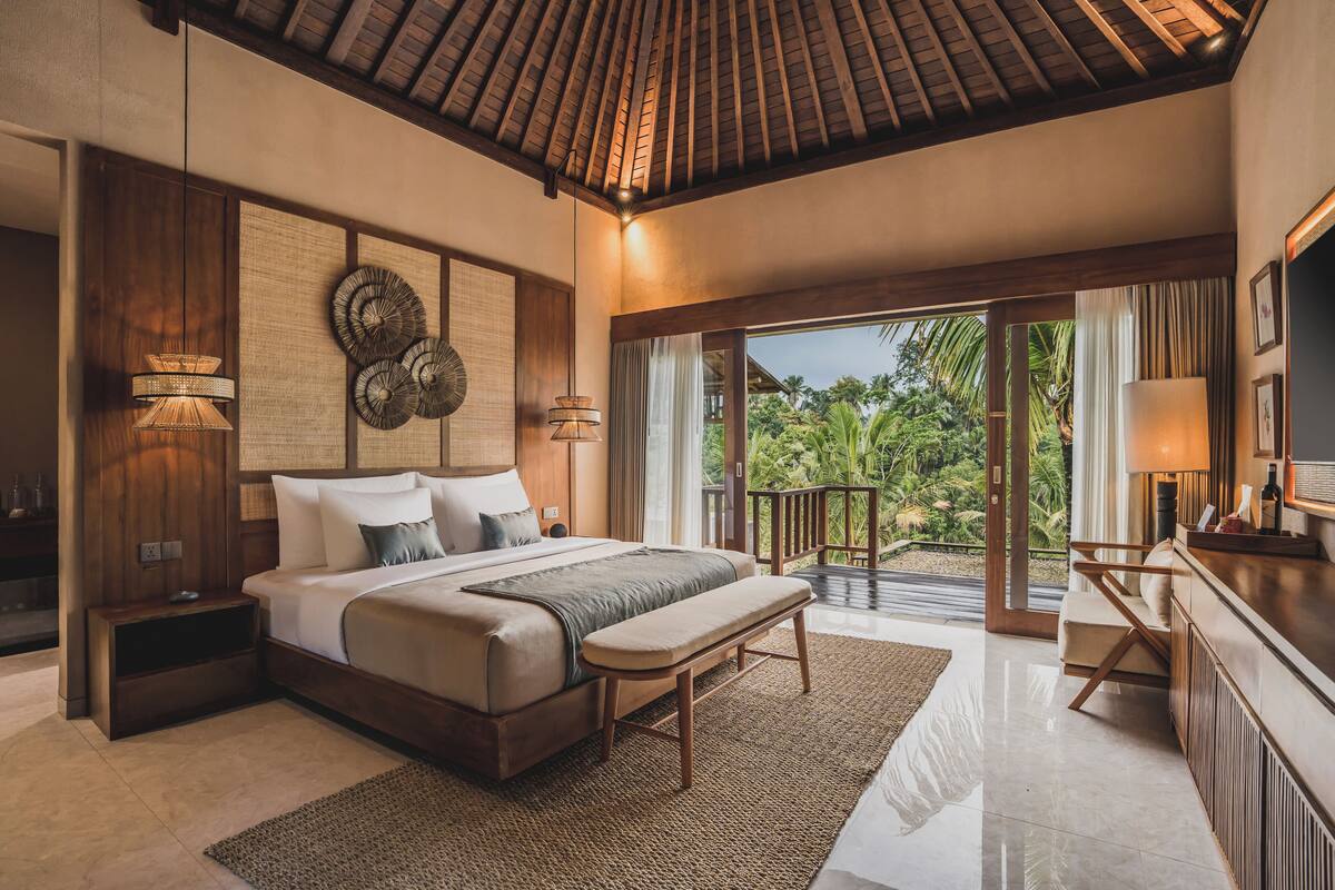 A spacious bedroom with a large bed, a bench, a desk, and a balcony with a view of greenery.