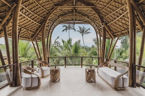 Habitación con estructura de bambú, dos sofás blancos acolchados y mesas redondas, con vistas a exuberante follaje y palmeras.