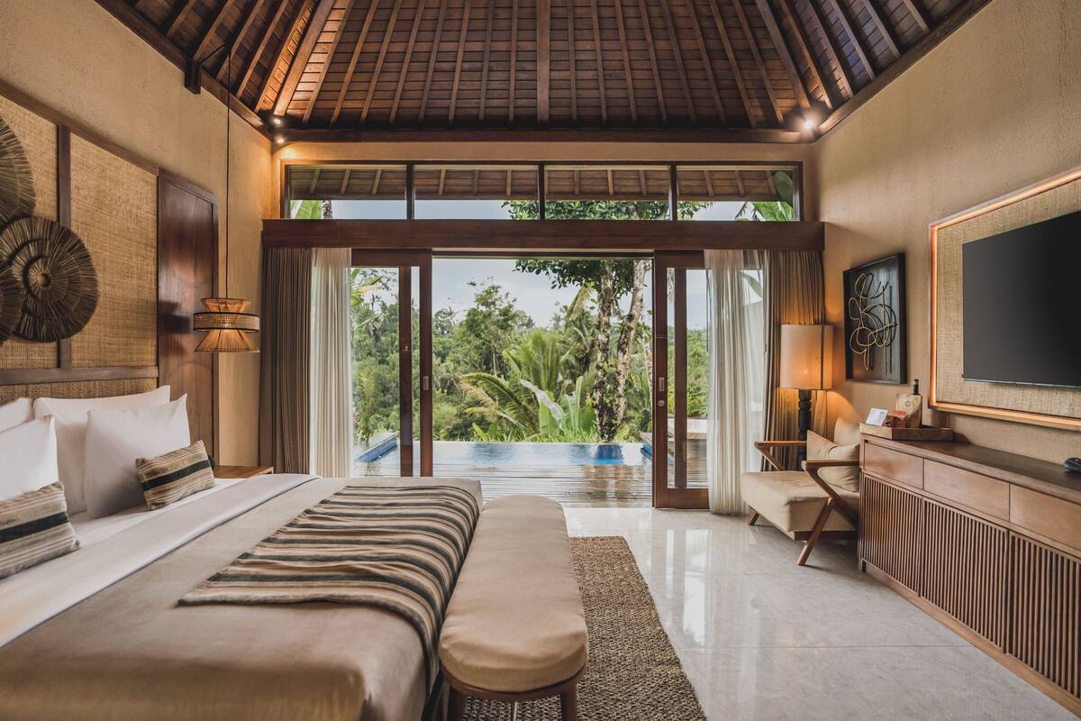 A spacious bedroom with a large bed, a bench, a desk, and a television. There is a sliding glass door leading to a balcony with a view of greenery.