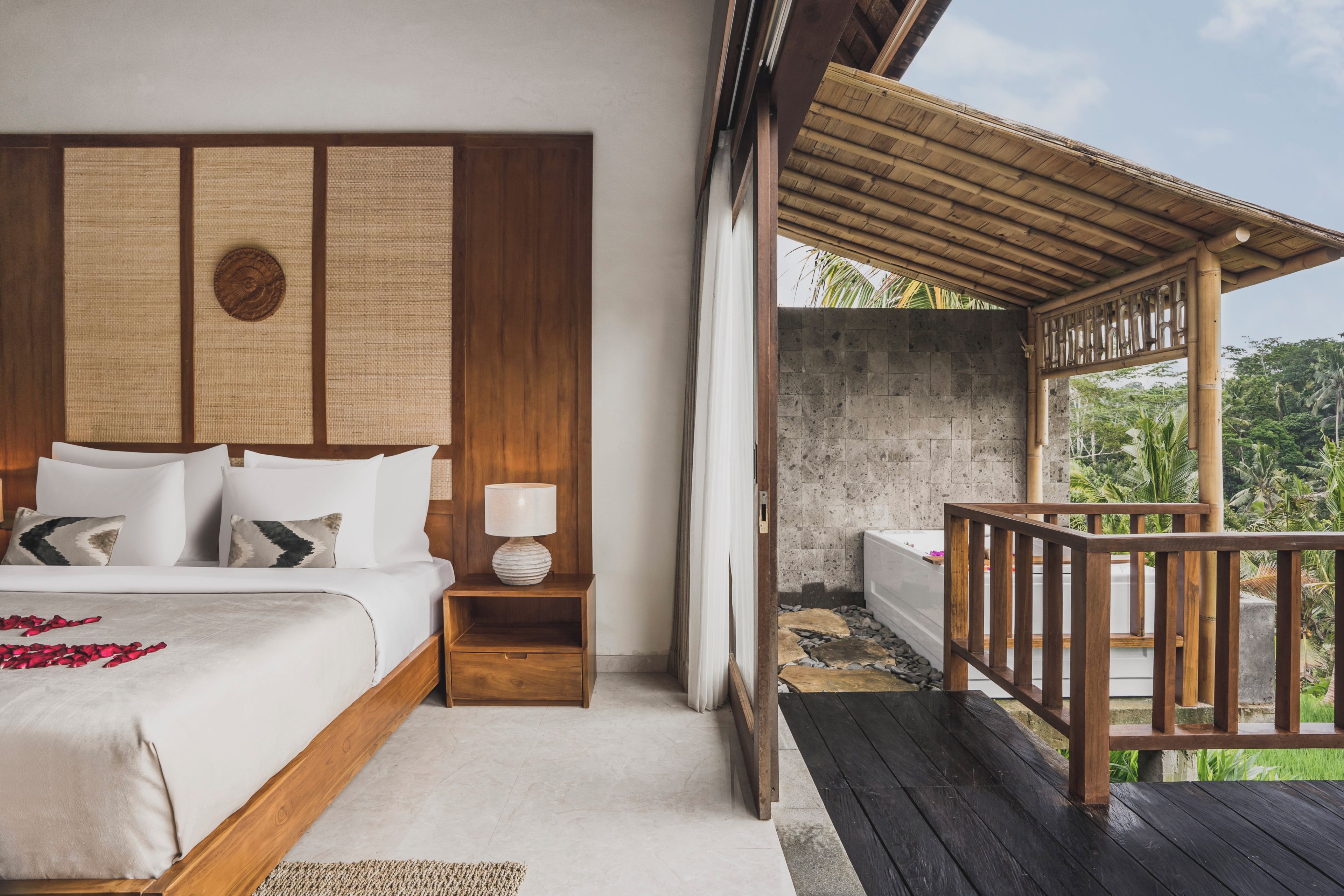A bedroom with a wooden headboard, a bed with white linens, a nightstand with a lamp, and a view of a balcony with a wooden railing.