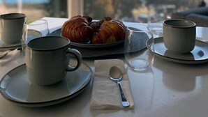 A table with a croissant, two cups, a spoon, and a napkin, with a view of a bridge and water.
