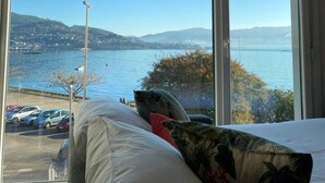 A bedroom with a large window offering a view of a body of water, a parking lot, and distant hills.