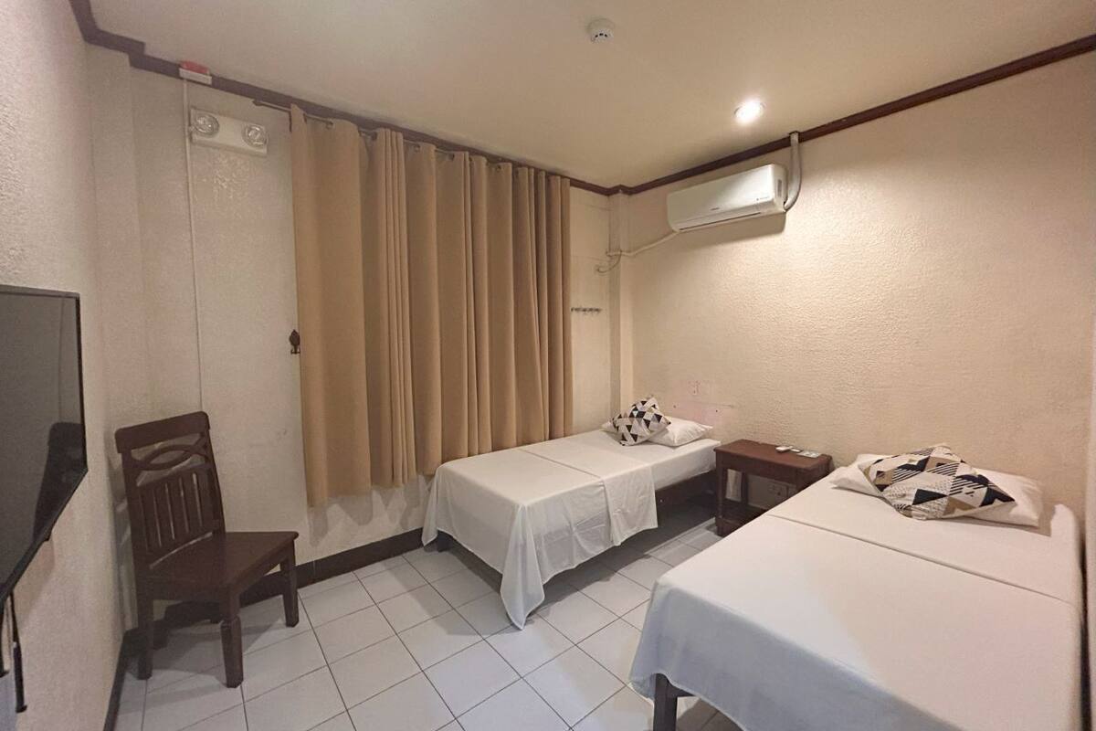 A hotel room with two beds, a chair, a TV, and an air conditioning unit.