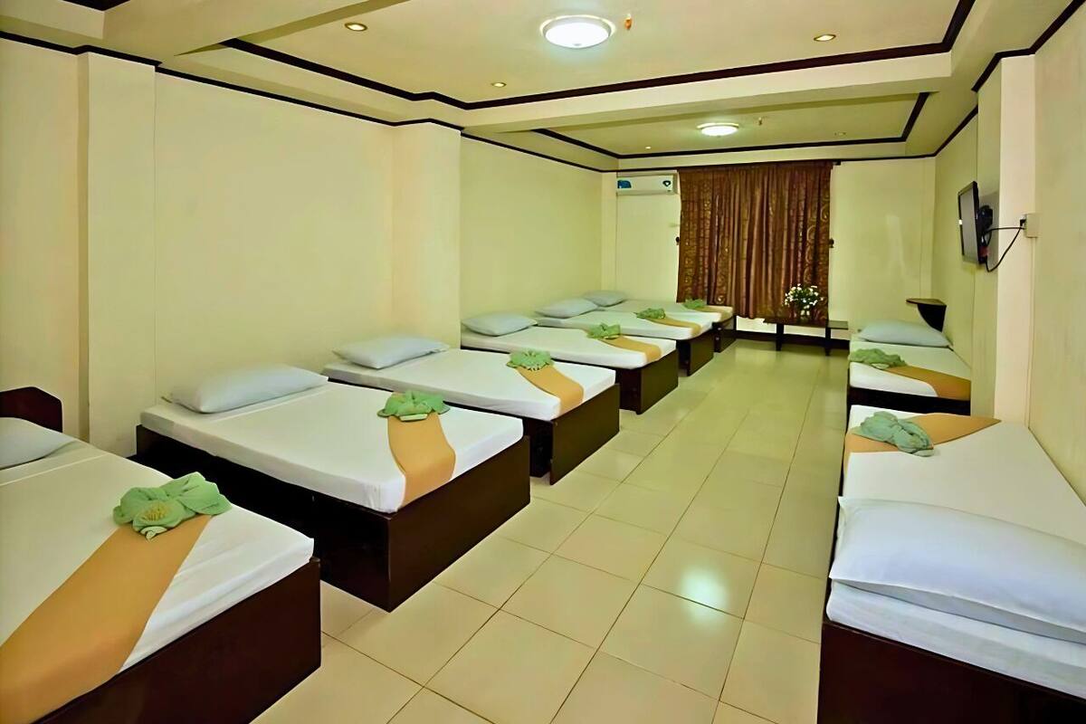 A hotel room with multiple beds arranged in a row, a ceiling with recessed lighting, and a visible air conditioning unit.