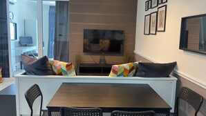 A modern living room with a dark wooden table, black perforated chairs, a large flat-screen TV, and colorful cushions.
