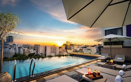 A rooftop pool area with lounge chairs and umbrellas, overlooking a cityscape at sunset.