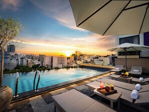A rooftop pool area with lounge chairs and umbrellas, overlooking a cityscape at sunset.