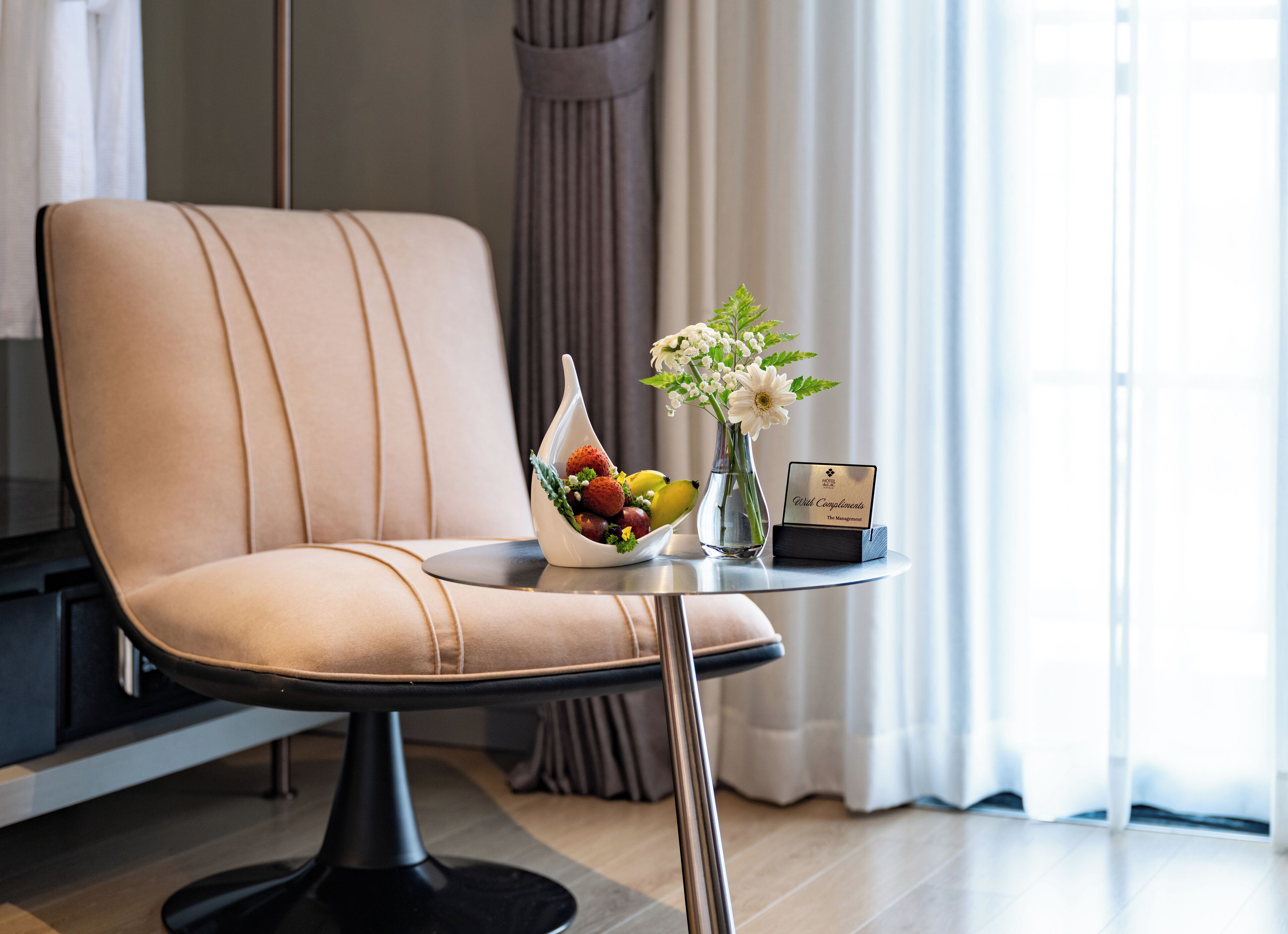 Et moderne hotelværelse med en behagelig stol, et lille bord med en skål frugt, en vase med blomster og et skilt med teksten "With Compliments The Management".