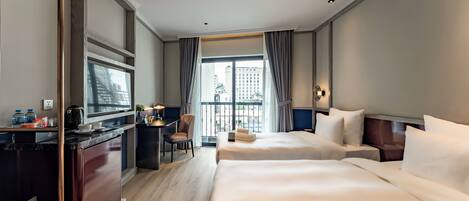 Habitación de hotel con dos camas, un escritorio con televisor y vistas a la ciudad a través de amplias ventanas.