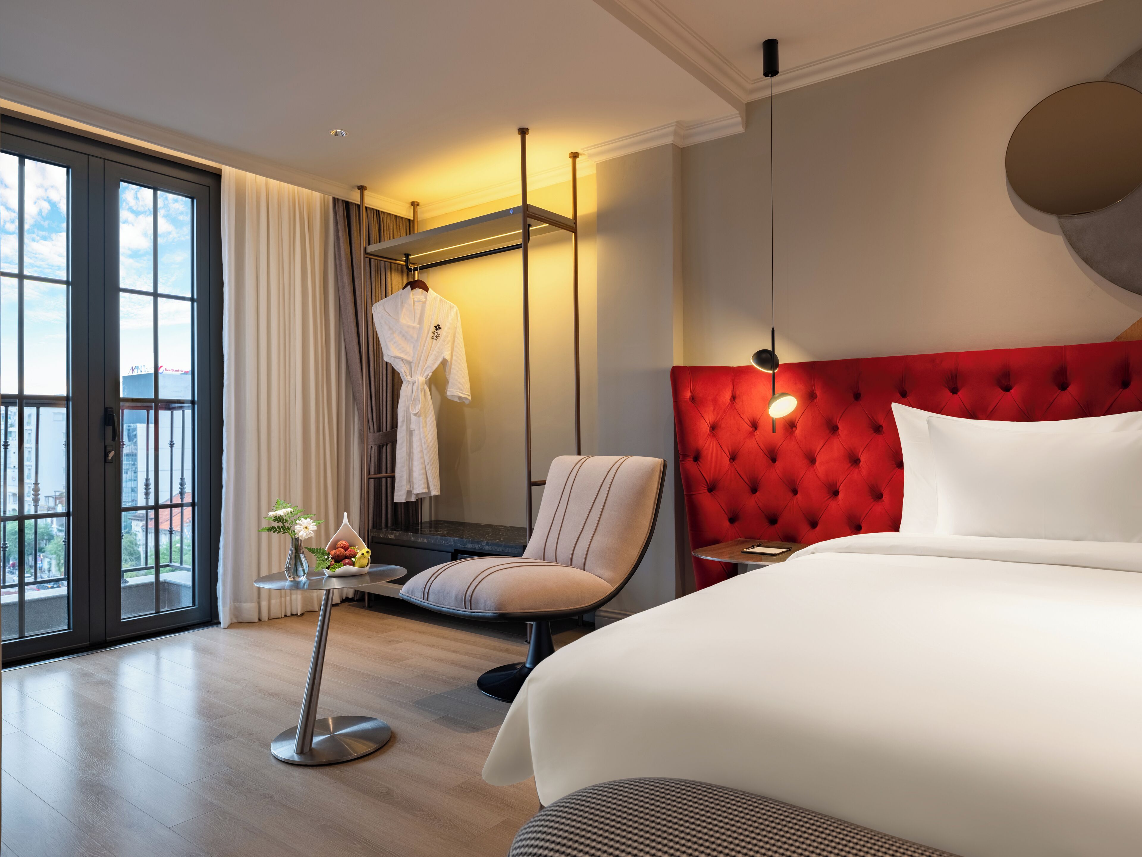 A hotel room with a large bed, a red upholstered headboard, a sitting area with a chair and a small table, a robe hanging on a rack, and a view through large windows.