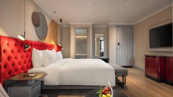 Una habitación de hotel moderna con una cama grande, cabecera tapizada en rojo, un televisor de pantalla plana, piso de madera y un frutero sobre la mesita de noche.