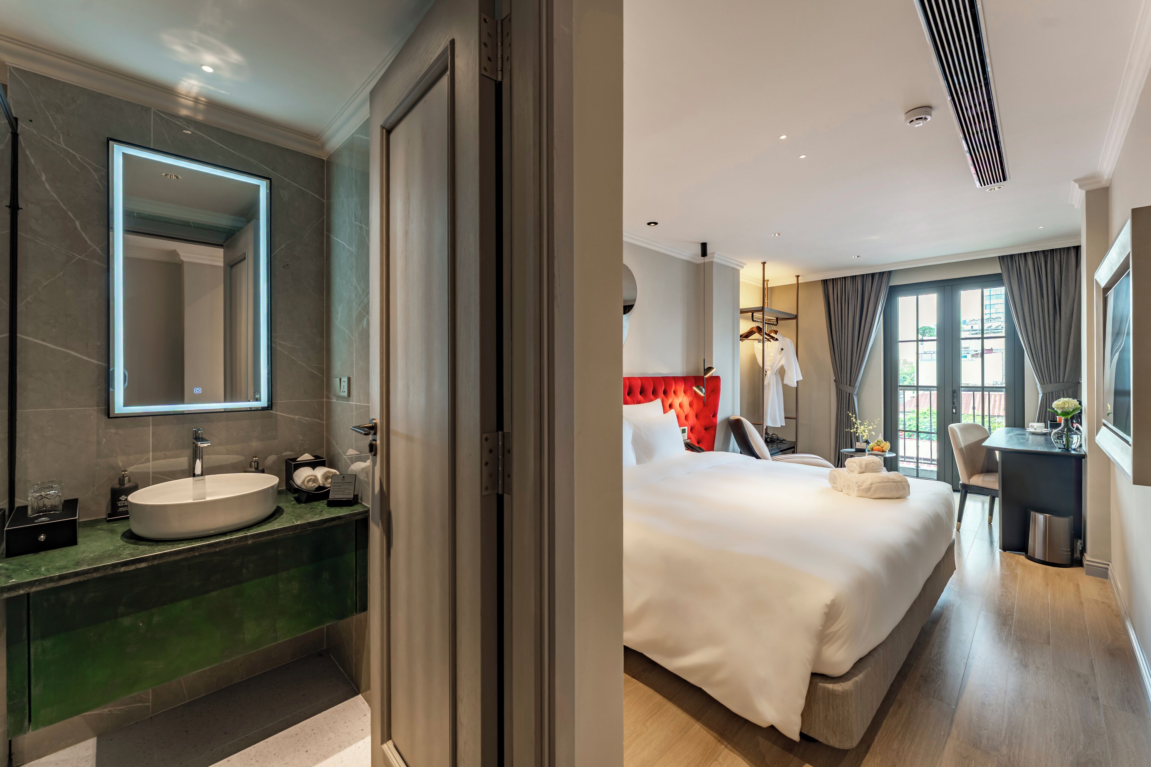A modern bathroom with a large mirror, sink, and green countertop. The room features a bed with white linens and a red headboard, a desk with a chair, and a window with curtains.