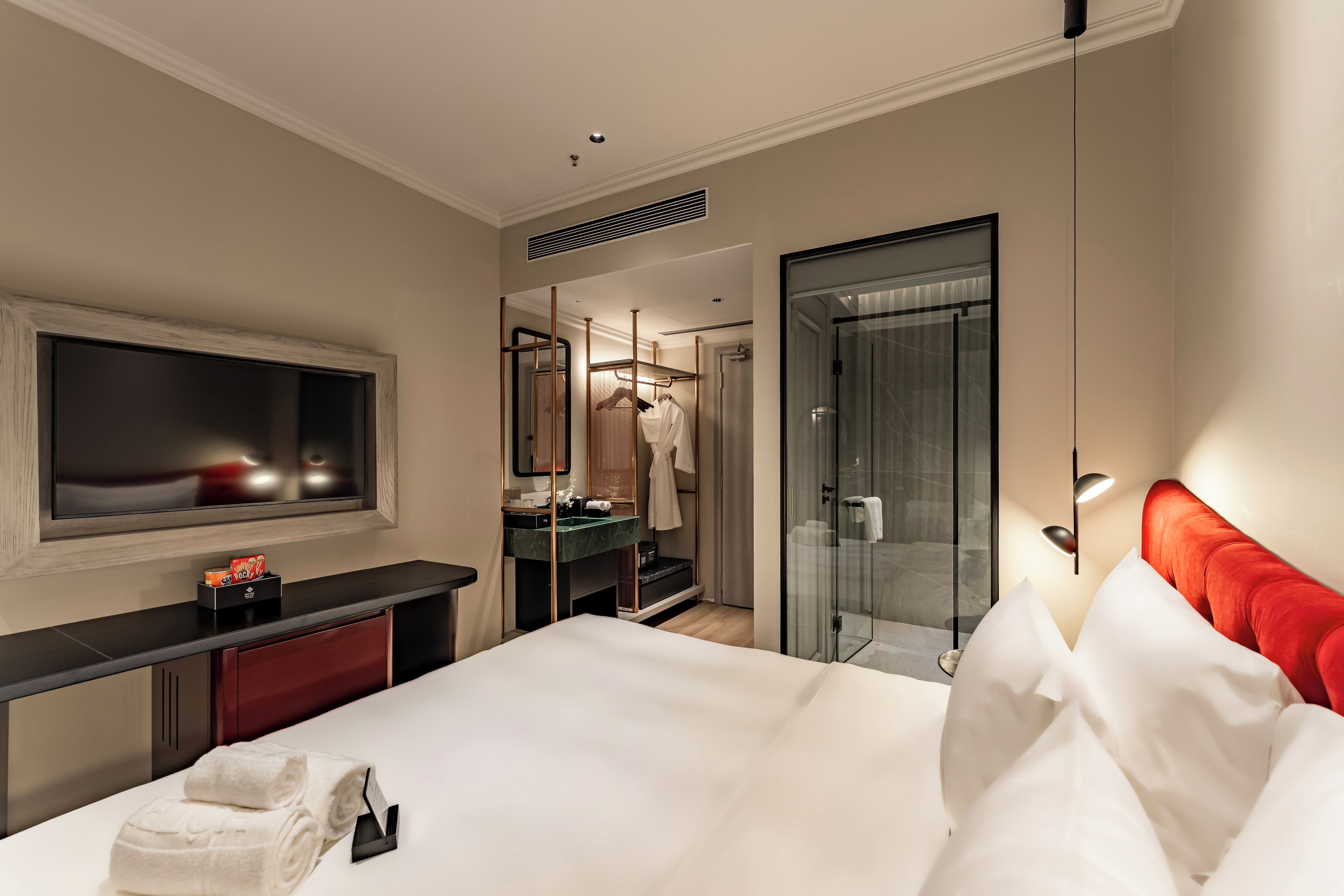 Habitación de hotel con cama, televisión, baño con espejo y cabecera roja.