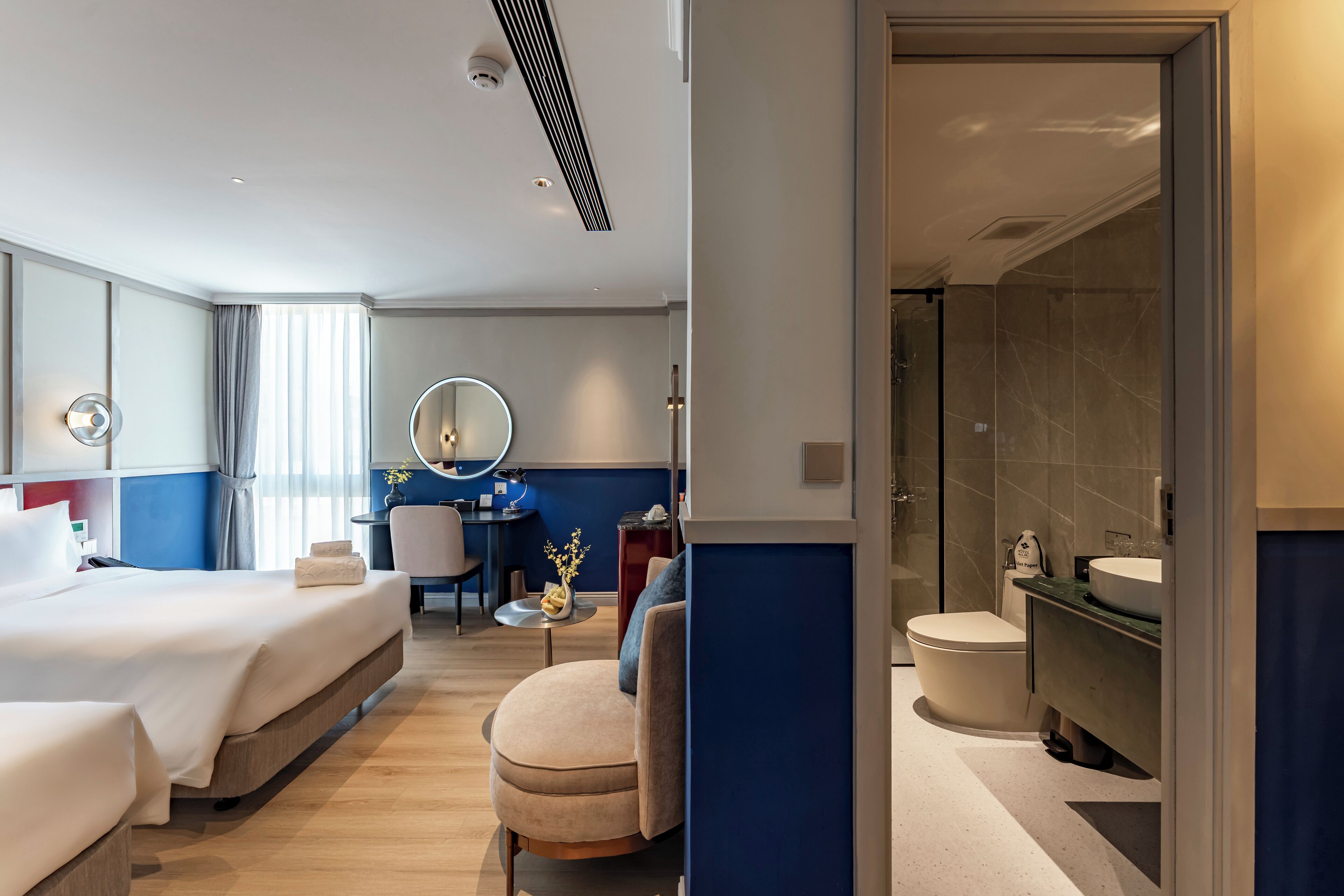A hotel room with a bed, a desk, a chair, a round mirror, a bathroom with a toilet and sink, and a shower area.