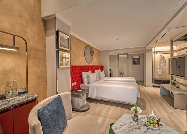 A hotel room with a red upholstered headboard, two beds, a TV, a minibar, and a small table with a vase and fruit.