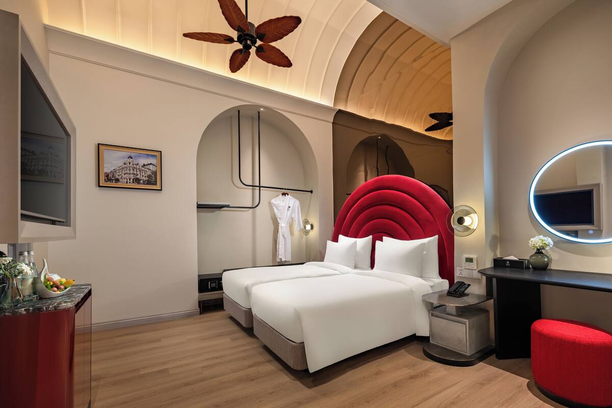 A modern hotel room with a large bed, a red accent wall, a round mirror, and a ceiling fan.