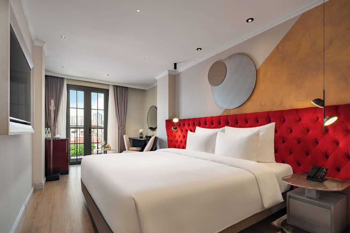 A modern hotel room with a large bed, a red tufted headboard, a bedside table with a lamp, and a view of the city through the window.