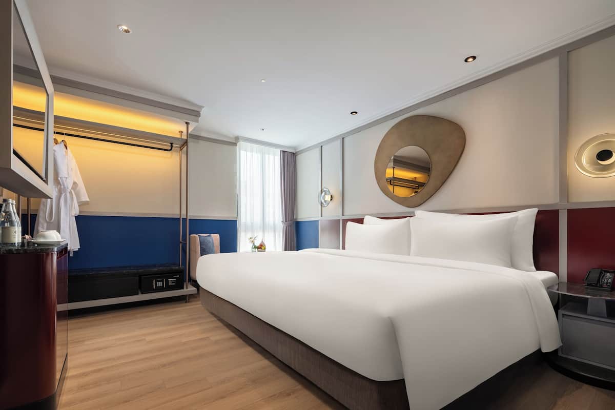 A modern hotel room with a large bed, a bedside table with a phone, a mirror, and a view of the cityscape through the window.