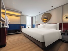 A modern hotel room with a large bed, a bedside table with a phone, a mirror, and a view of the cityscape through the window.
