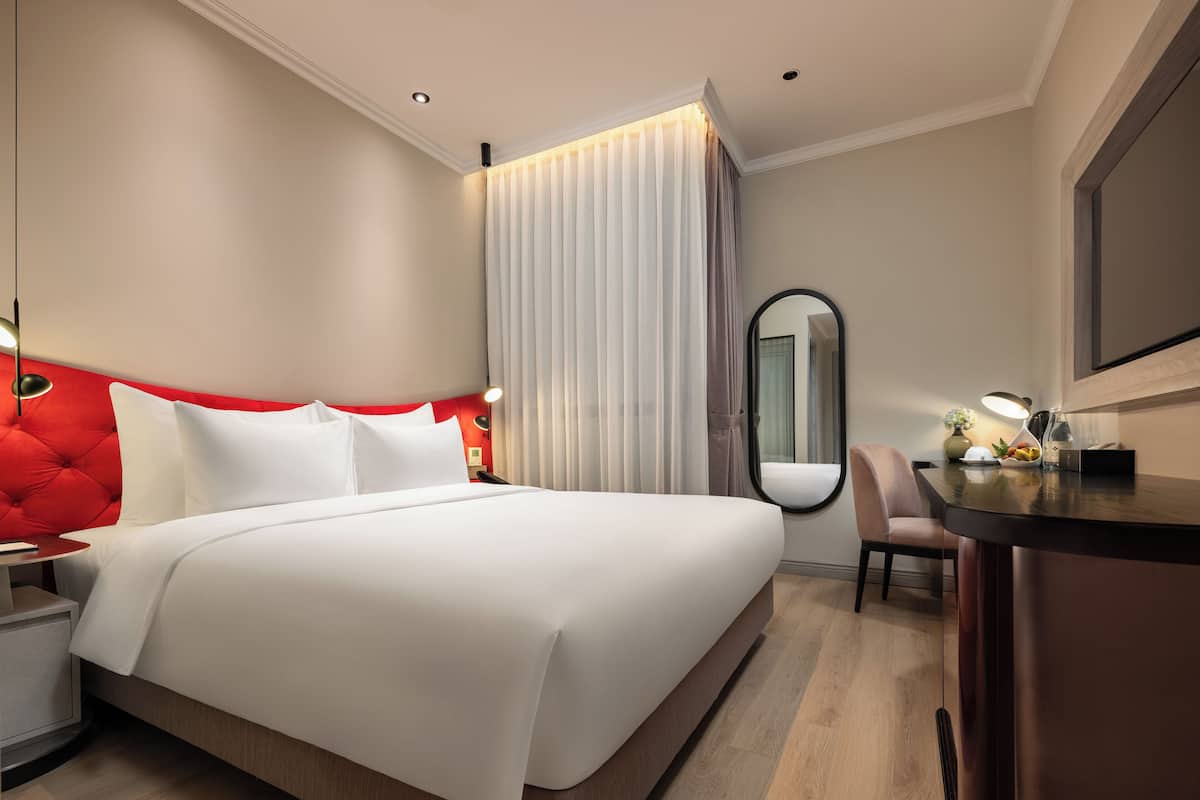 A modern hotel room with a large bed, a desk, a chair, and a mirror.
