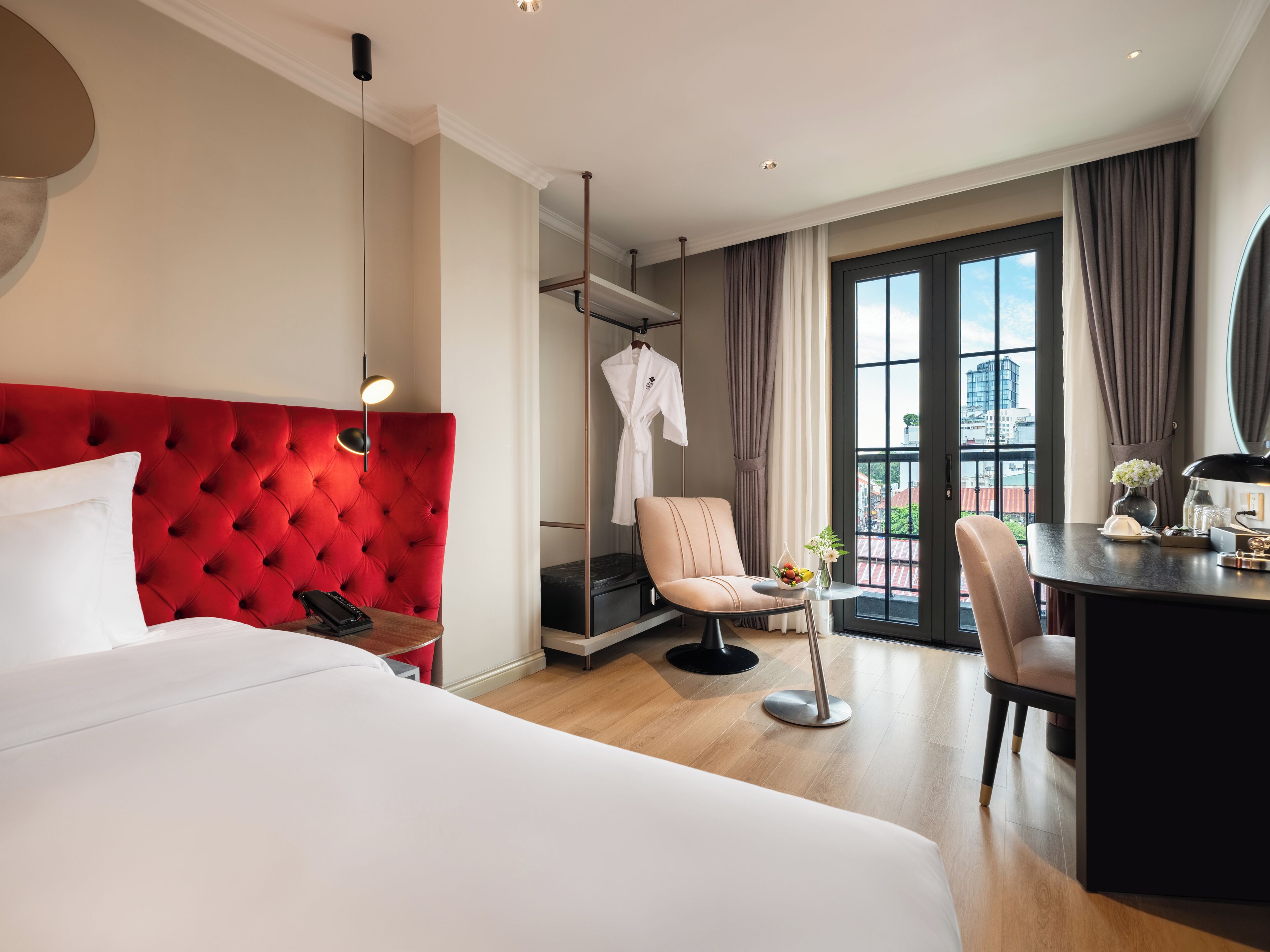 Ein Hotelzimmer mit einem roten, gepolsterten Kopfteil, einem Bett, einem Schreibtisch mit Stuhl, einem kleinen Tisch mit Blumenvase und Blick auf Gebäude durch große Fenster.