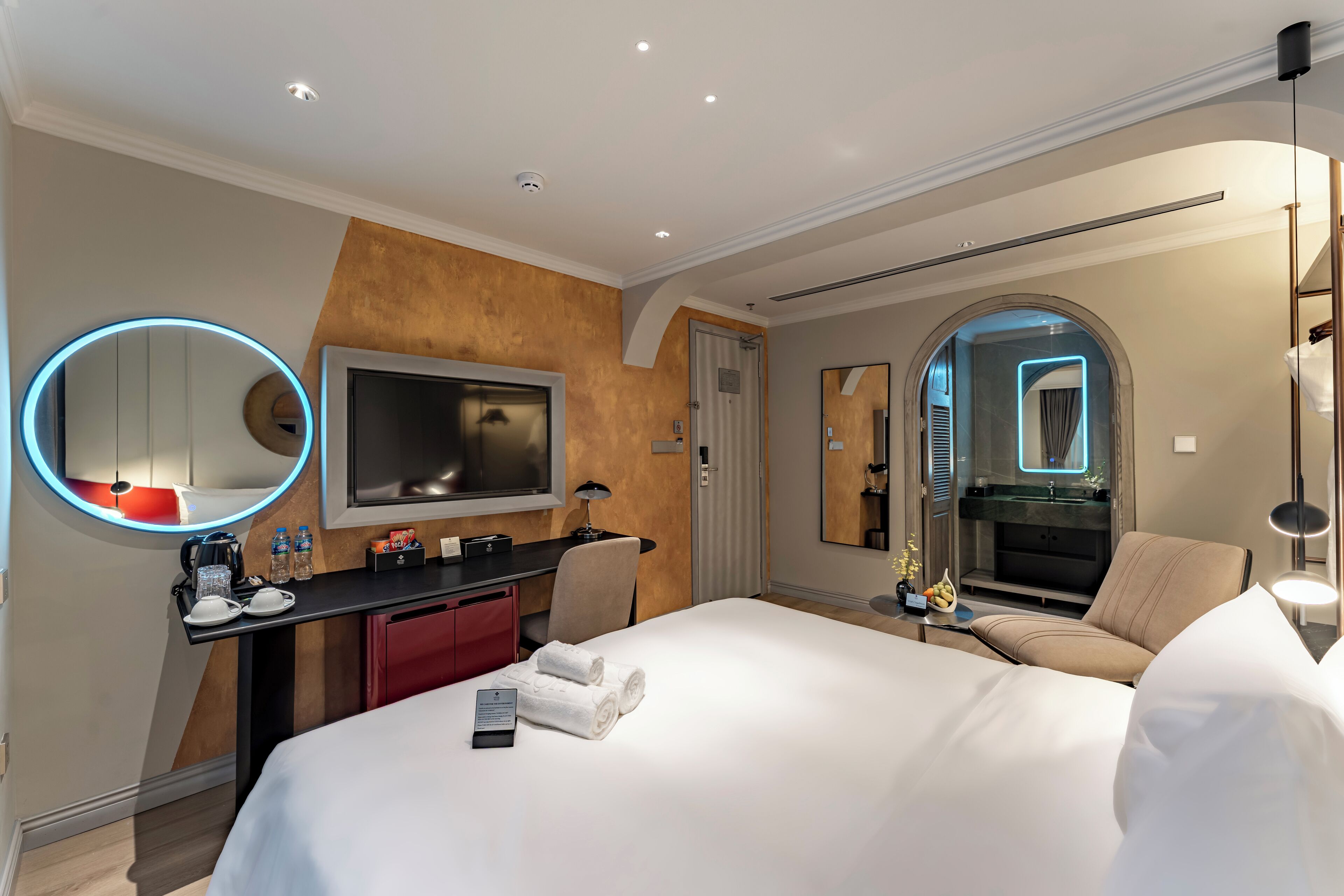 A modern hotel room with a large bed, a desk with a flat-screen TV, a round mirror with a neon light, and a bathroom with a glass shower.