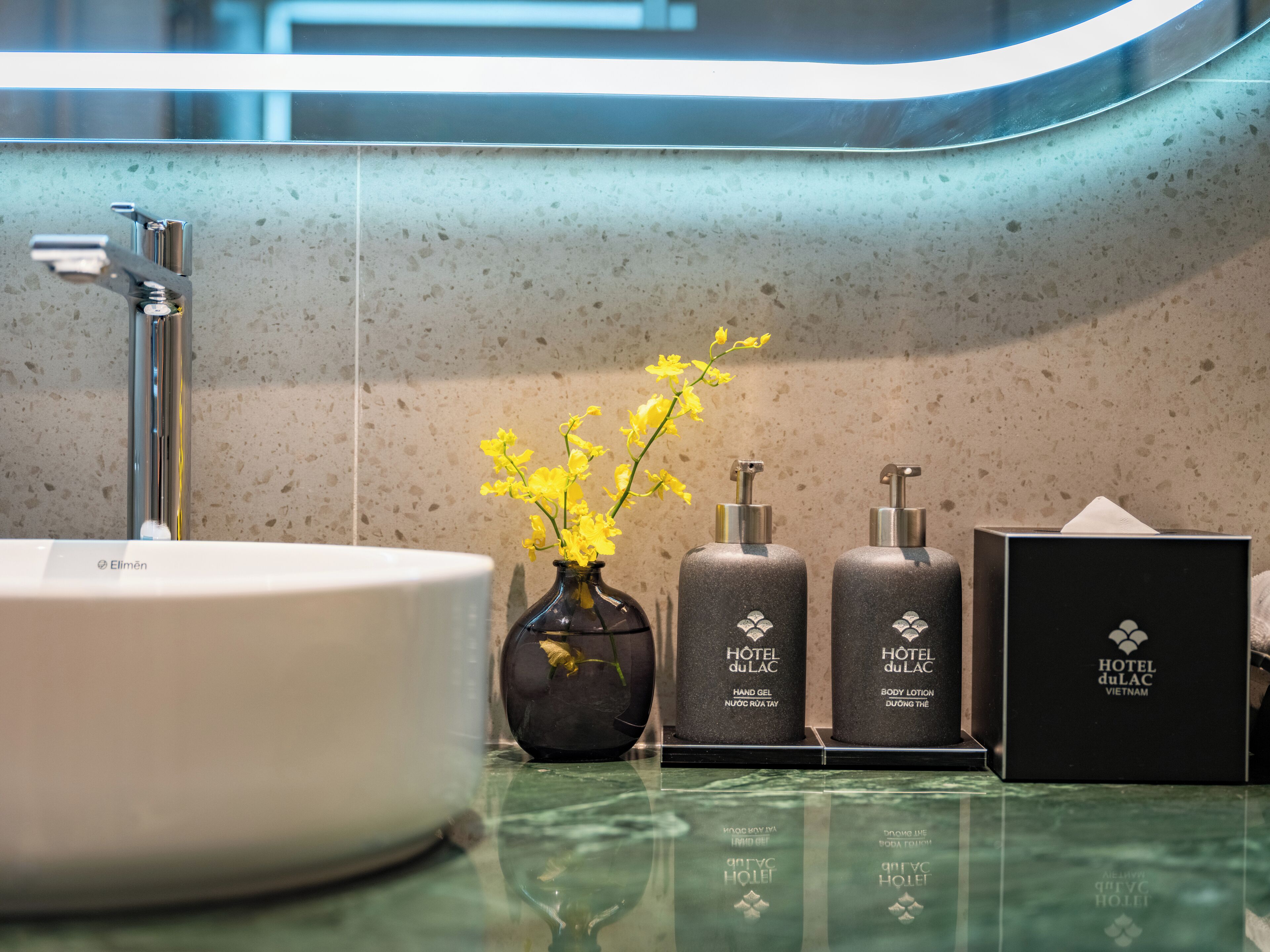 A bathroom with a round sink, a mirror, a vase with yellow flowers, and two bottles of hotel lotion.