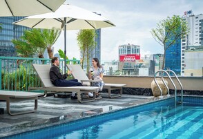 A rooftop pool area with lounge chairs and umbrellas, surrounded by greenery and city buildings.