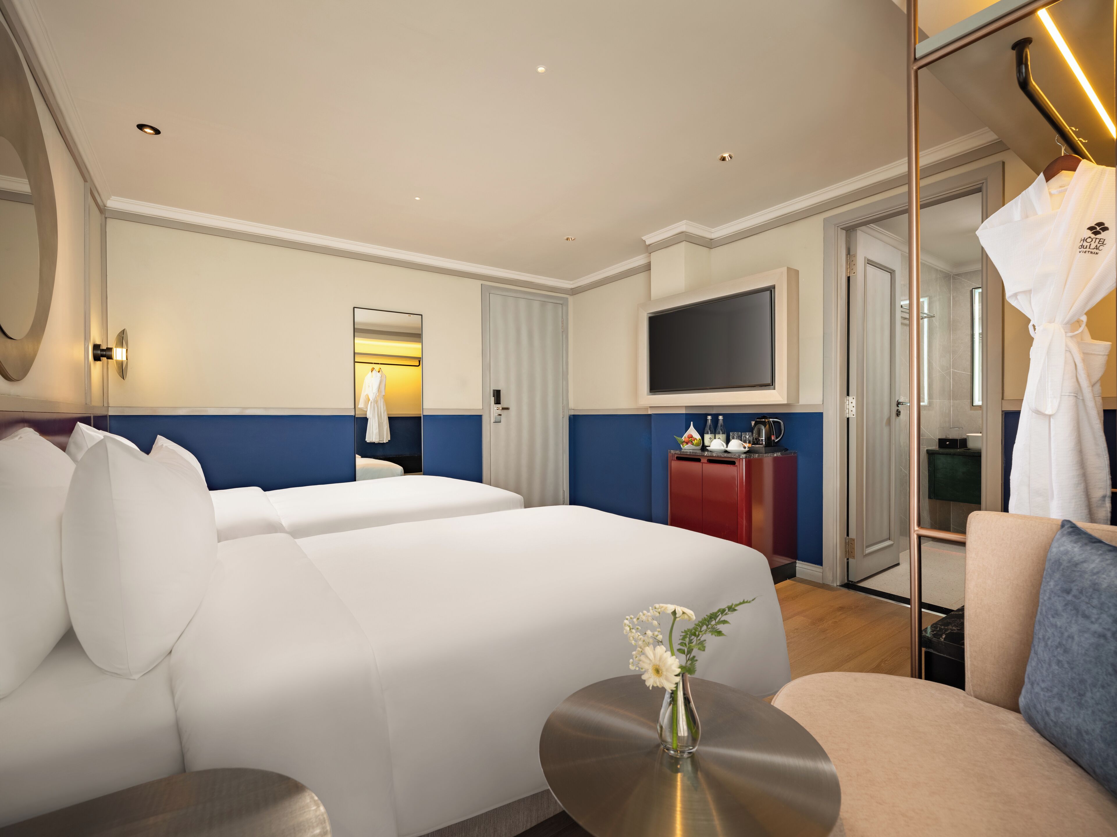 A hotel room with two beds, a TV, a minibar, and a bathroom visible through a glass door.