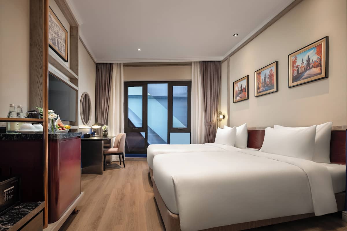 A hotel room with a large bed, a desk with a computer, a chair, a window with curtains, and framed pictures on the wall.