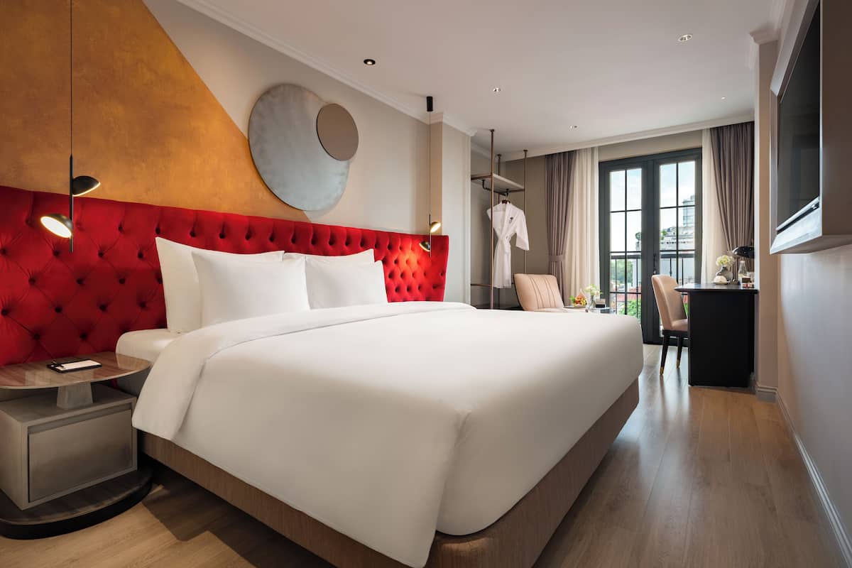 A modern hotel room with a large bed, a red upholstered headboard, a round wall decoration, a bedside table, and a view of the city through large windows.