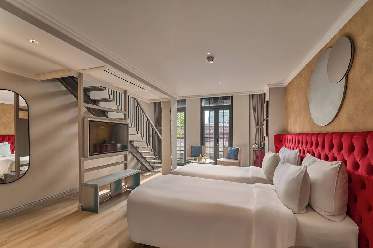A hotel room with a large bed, a red upholstered headboard, a TV, a staircase, and a view of the outside.