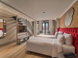 Habitación de hotel con una cama grande, un cabecero tapizado en rojo, un televisor, una escalera y vistas al exterior.