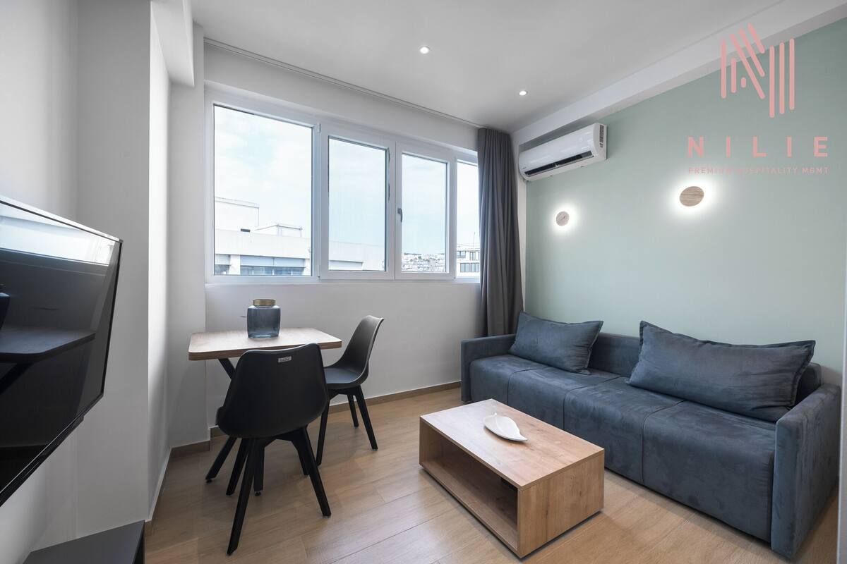 Ein moderner, gut beleuchteter Raum mit einem grauen Sofa, einem Holztisch, einem kleinen Tisch mit Stuhl und einem Fernseher.