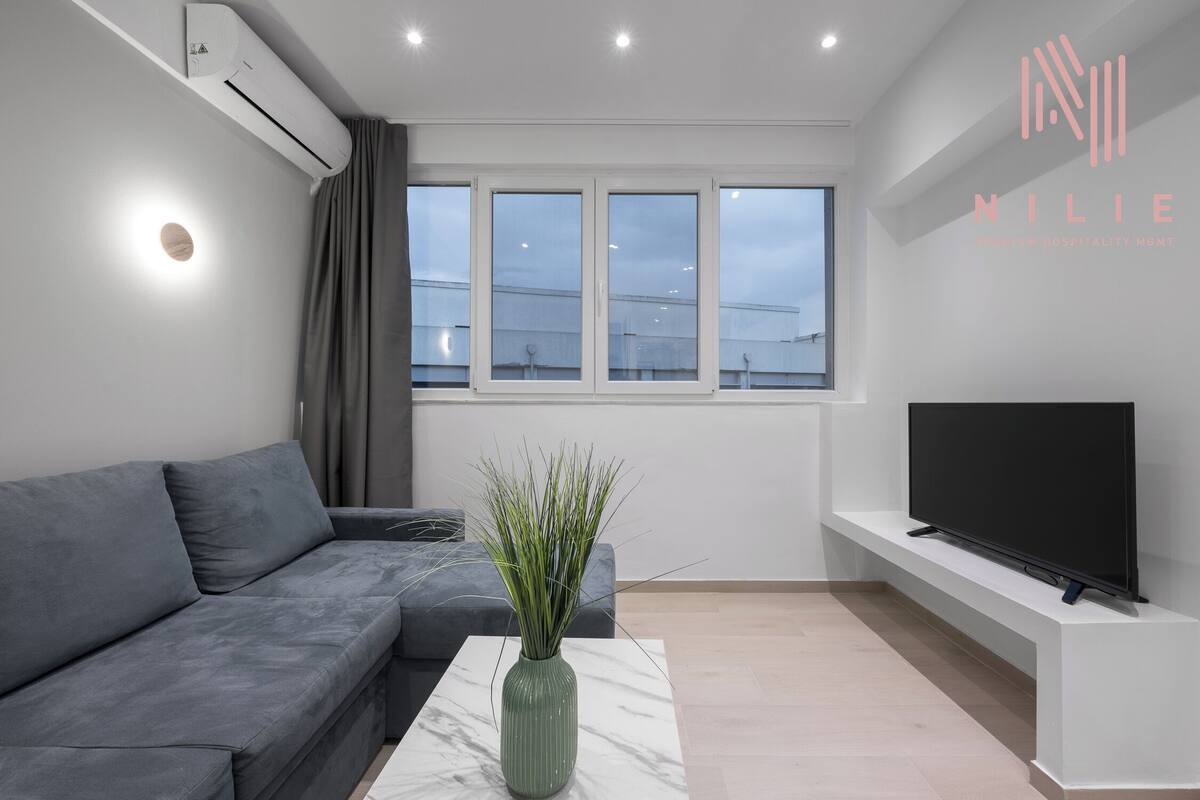 Ein modernes Wohnzimmer mit einem grauen Sofa, einem мраморном Couchtisch, einer grünen Vase mit Pflanzen, einem Flachbildfernseher auf einem weißen Schrank und großen Fenstern mit Stadtblick.