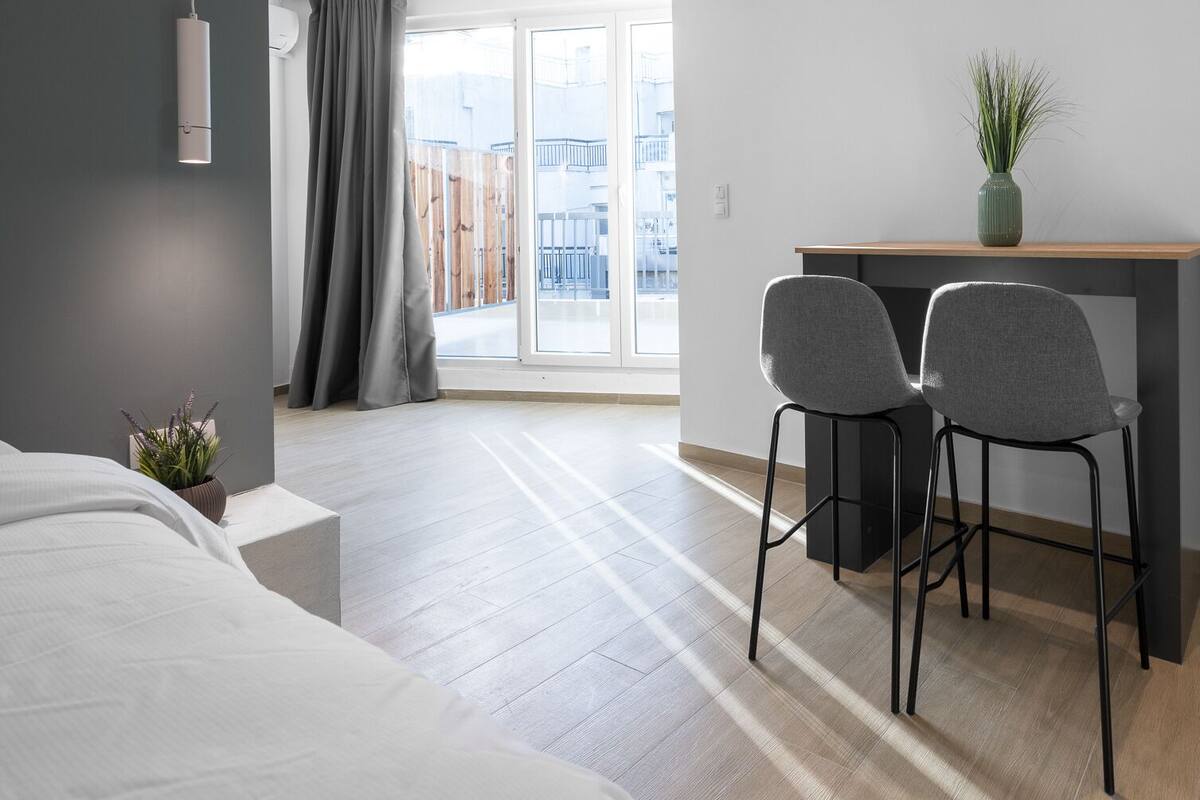 Ein modernes Hotelzimmer mit einem Bett, einem Schreibtisch mit zwei Stühlen, einer Zimmerpflanze und Blick auf Gebäude durch große Fenster.